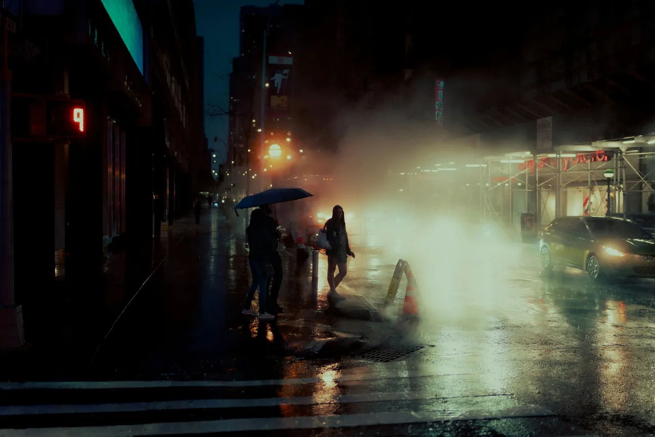 A rainy city street at night, steam rising from the pavement as people pass under umbrellas.