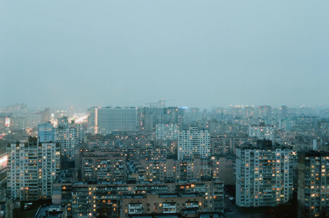 A dense city of apartment towers at dusk, windows starting to light up.