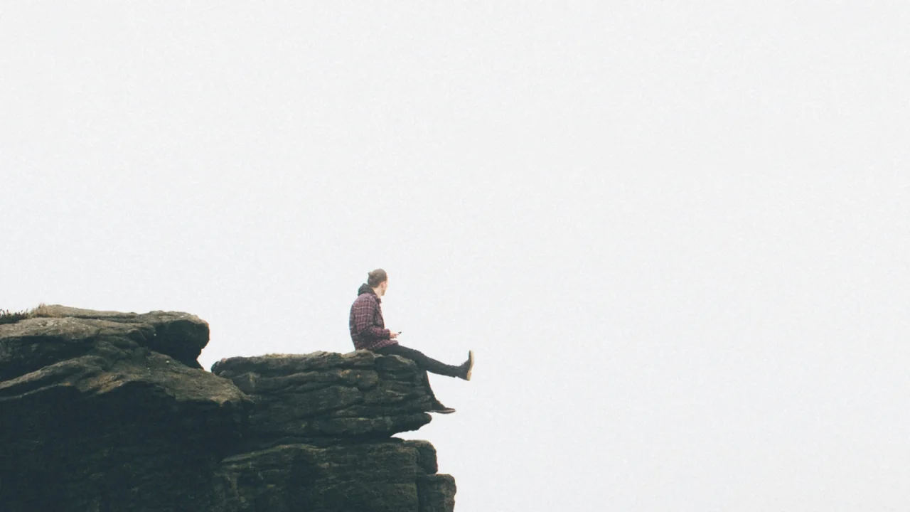 A man sits on the edge of a rock outcrop, legs out over the drop, looking into thick fog.