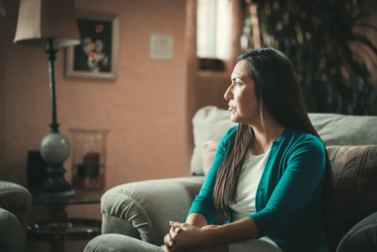 A woman sits on a couch in a dim living room, looking out a window.